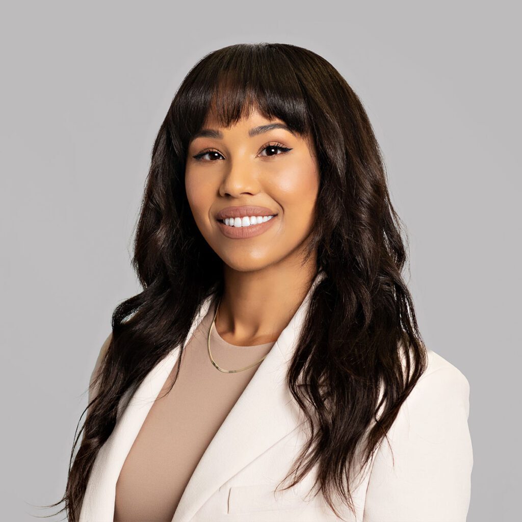 A person with long dark brown hair smiles a the camera. They are wearing a white blazer with a beige blouse underneath and a silver necklace.